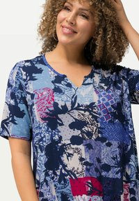 Krátký rukáv tunika v modré barvě s patchworkovými květinovými vzory, s výstřihem do V a různými texturami v námořnické, červené a bílé akcenty.