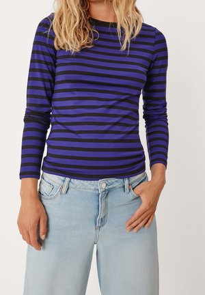 Femme portant une chemise à manches longues rayée horizontalement bleu et noir, associée à un jean taille haute bleu clair, debout devant un fond uni.