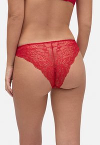 Tanga de encaje rojo con patrón floral, que presenta detalles intrincados y una textura transparente. La cinturilla elástica se coloca por debajo de las caderas.