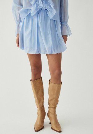 Woman wearing a light blue ruched mini dress and tan knee-high suede boots, standing against a plain light background.