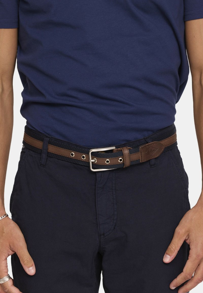 Man in een marineblauwe broek en shirt met een bruine leren en zwarte stoffen riem om de taille gestrikt.