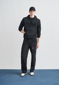 Felpa nera con logo Adidas testurizzato, abbinata a jeans neri strappati con borchie decorative. Il modello indossa anche un berretto nero e sneakers.