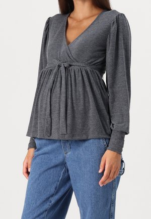 Femme portant un top cache-cœur à manches longues gris foncé avec un lien à l'avant et un jean en denim bleu, montrée du cou aux hanches.