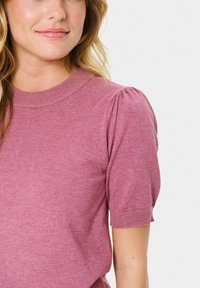 Frau mit blonden Haaren, die ein pinkes, kurzärmeliges Strickoberteil mit gerafften Schultern und einem gerippten Rundhalsausschnitt sowie Bündchen trägt.