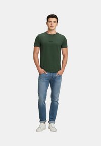 T-shirt verde a maniche corte con scollatura rotonda e branding sottile; abbinato a jeans azzurri chiari e sneakers bianche.