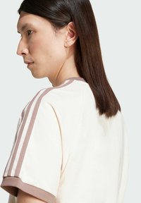 Κρέμα μπλούζα με στρογγυλή λαιμόκοψη, κοντά μανίκια που διαθέτουν δύο κάθετες καστανές ρίγες και υφή υφάσματος.