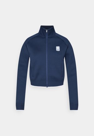 Chaqueta azul marino con cremallera, cuello alto, mangas largas y un logo blanco en la parte superior izquierda del pecho. Presenta un diseño ajustado y tela texturizada.