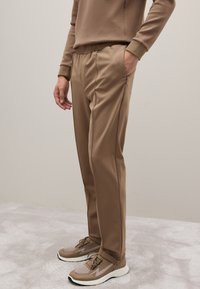 Beige Jogginghosen mit einem Kordelzugbund, schmal zulaufenden Beinen und seitlichen Taschen, kombiniert mit hellbraunen Sportschuhen mit Mesh-Akzenten.