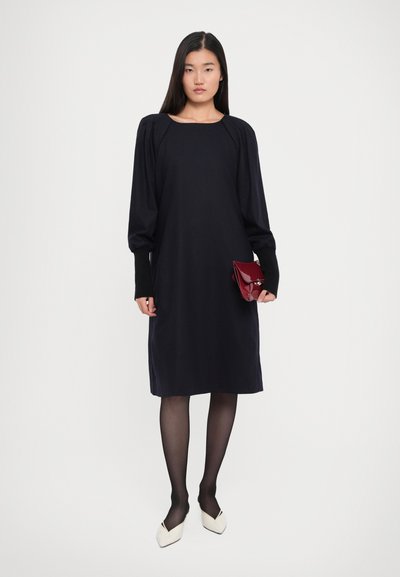 Woman wearing a black long-sleeve midi dress, sheer black tights, white pointed shoes, and holding a small red handbag against a plain background.