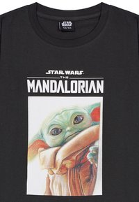 Schwarzes T-Shirt mit dem Text "Star Wars The Mandalorian" über einer detaillierten Illustration von Baby Yoda, der in einem beigen Umhang gehüllt ist.