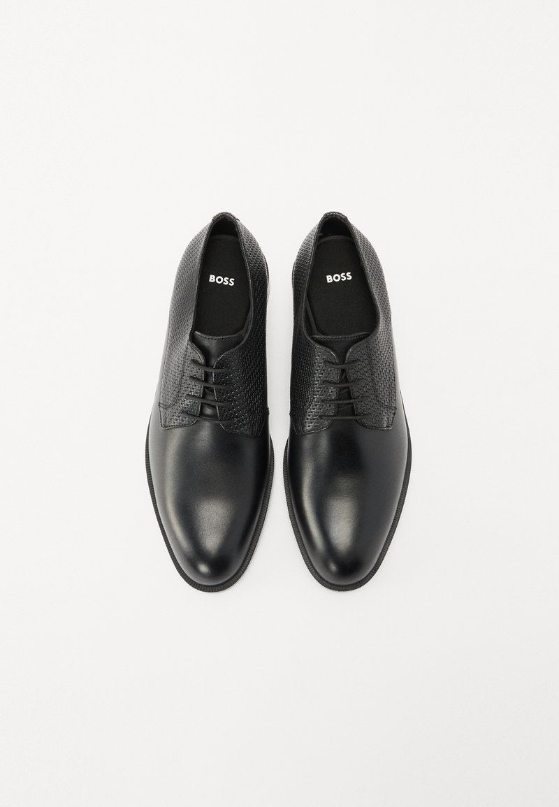 Chaussures en cuir noir avec une finition lisse et des panneaux en mesh texturé. Comprend des lacets et un logo sur les semelles intérieures. Bout arrondi et design minimaliste.