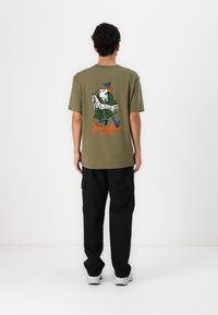 Olivově zelené bavlněné tričko s kresleným designem na zadní straně, se postavou držící mapu a textem "Barbour" v oranžové barvě.