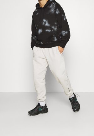 Personne portant un sweat à capuche tie-dye noir et blanc, un pantalon de jogging écru et des baskets noires, debout avec une jambe en avant et les mains dans les poches.