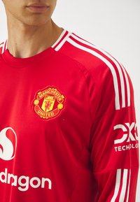 Raudona „Manchester United“ marškiniai su baltomis juostelėmis ant pečių. Yra tekstūruotas audinys, išsiuvinėtas logotipas ir prekės ženklas ant rankovės.