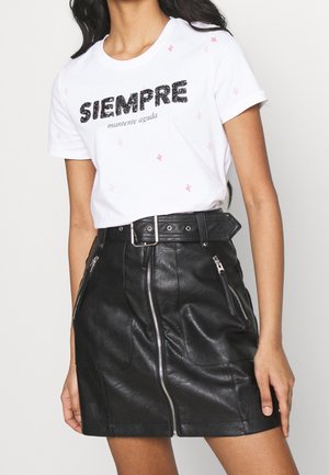 Femme portant un T-shirt blanc avec le texte "SIEMPRE mantente aguda" et une jupe en cuir noire avec une fermeture éclair à l'avant et une ceinture.