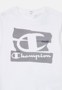 Hvid bomulds T-shirt med et sort mesh-mønsteret Champion-logo på forsiden, rund halsudskæring og trykknaplukning bagpå.