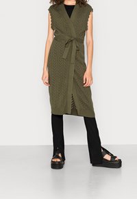 Olivgrön stickad ärmlös cardigan med V-hals, vågiga kanter och band i midjan, matchad med svarta byxor med delad nederkant och klumpiga sandaler.