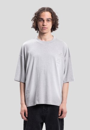 Jeune personne aux cheveux bouclés portant un t-shirt ample gris clair et un pantalon foncé, debout devant un fond uni clair.