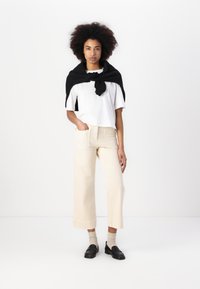 Top corto bianco a maniche corte, pantaloni beige a gamba larga, maglione nero appoggiato sulle spalle, calze beige, mocassini neri su uno sfondo neutro.