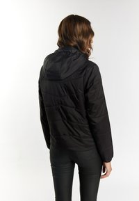 Zwarte pufferjas met een capuchon, gequilt textuur, elastische manchetten en een slim-fit silhouet, gedragen met zwarte strakke broek.