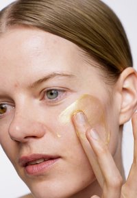 Crema dorada siendo aplicada sobre la piel con un dedo; textura suave, consistencia semitranslúcida, resaltando los detalles faciales.