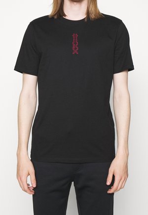 Personne portant un t-shirt noir uni à manches courtes avec un petit motif abstrait vertical rouge au centre de la poitrine.