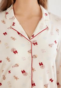 Koszula piżamowa w kremowym materiale z czerwonymi lamówkami, zdobiona wzorem z piernikami, lizakami, kokardkami i deserami. Guziki przy kołnierzyku.