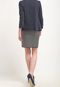 Marinblå kavaj med trekvartsärmar, kombinerad med en svartvit randig pennkjol. Modell som bär marinblå klackar, sedd bakifrån.