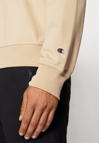 Beige Baumwoll-Sweatshirt mit gerippten Bündchen. Verfügt über ein kleines gesticktes Logo in Rot, Weiß und Blau an einem Bündchen. Hand ruht auf schwarzem Stoff.