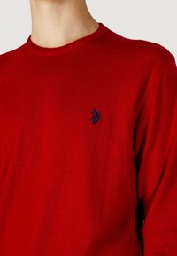 Maglione rosso a maglia con scollo rotondo, caratterizzato da un piccolo logo blu navy sul petto. La trama è liscia e morbida.
