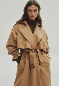 Cappotto trench color khaki con cintura, ampi rever e dettagli a bottoni. Presenta maniche arricciate e una vestibilità morbida. Tessuto liscio, senza motivi.