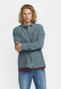 Overhemd met knopen in teal stof met een gestructureerde afwerking, voorzien van een borstzak en bruine knopen. Gelaagd over een bordeauxrood shirt.