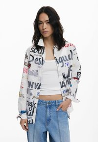 White button-up shirt featuring multicolored printed text and graphics. Long sleeves, relaxed fit, paired with light blue denim jeans.