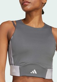 Gray athletic crop top with thin shoulder straps, elastic waistband, and white branding accents. Smooth texture, fitted design, and minimalist style.