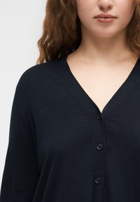 Νέα γυναίκα με καστανά σγουρά μαλλιά, που φοράει μαύρη μπλούζα με κουμπιά και V-λαιμόκοψη, εμφανίζεται από τους ώμους ως το πηγούνι πάνω σε λευκό φόντο.