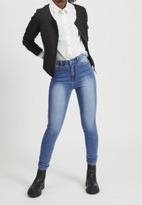 Svart texturerad jacka över en vit skjorta med knappar, kombinerad med blå skinny jeans och svarta ankelskor. Enkelt bakgrund.