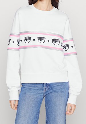 Vrouw draagt een witte trui met roze en zilveren strepen met zwarte oog- en sterrenpatronen, gecombineerd met blauwe spijkerbroek.