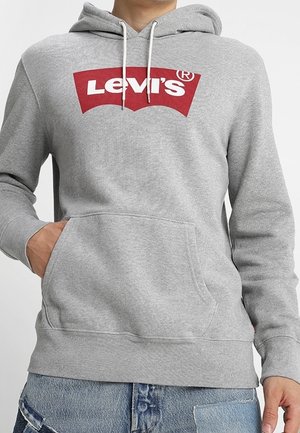 Osoba ubrana w szarą bluzę z kapturem Levi's z czerwonym logo, ręce w przedniej kieszeni, w połączeniu z niebieskimi dżinsami patchwork.