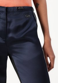Navy satin athletic pants with black side panels. Features a drawstring waist and a small logo accent on the front. Smooth texture.