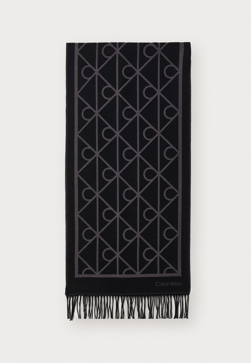 Μαύρο μαντήλι με γκρι γεωμετρικό σχέδιο, με φουντωτές άκρες. Το υλικό φαίνεται μαλακό και υφίσταται. Το λογότυπο της μάρκας είναι ορατό στην γωνία.