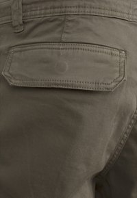 Los pantalones de color verde olivo oscuro tienen un tejido texturizado, un bolsillo lateral y un bolsillo trasero rectangular con costura en contraste.