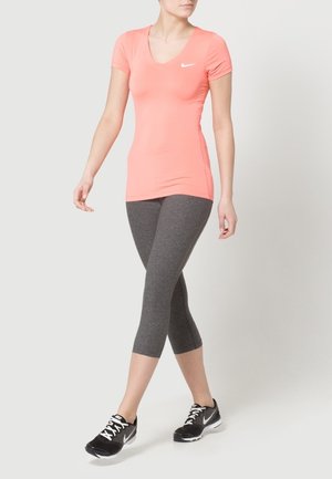 Kobieta ubrana w koralowy top Nike z krótkim rękawem, szare krótkie legginsy oraz czarne sneakersy Nike, stojąca z jedną stopą lekko wysuniętą do przodu.