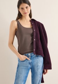 Débardeur marron en tissu lisse, associé à un cardigan bordeaux en tissu pelucheux. Jean bleu clair à taille haute avec fermeture à deux boutons.