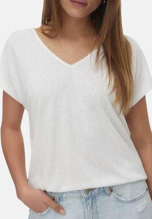 Vrouw draagt een witte V-hals T-shirt met gouden bies en lichtblauwe denim jeans, met één hand in de zak en steil bruin haar.