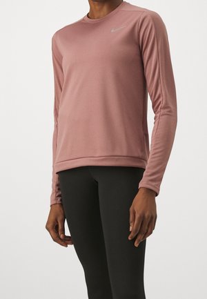 Personne portant un haut de sport mauve à manches longues avec logo Nike et un legging noir sur fond uni.