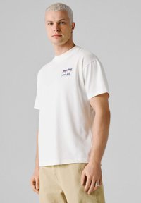 Mladý muž s krátkymi blond vlasmi, oblečený v bielom tričku s grafikou Red Bull Racing World Tour a béžových nohaviciach, stojaci na jednoduchom sivom pozadí.