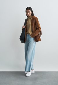 Brun suedjakke med frynse detaljer, beige høyhalset genser, lys blå vide jeans og hvite joggesko, kombinert med en svart veske.