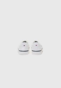 White canvas slip-on shoes with a low profile, featuring a black stripe along the sole and a small colored logo near the heel.