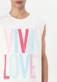 Hvit ermeløs skjorte med teksten "VIVA LOVE" i store, fete bokstaver med rosa, rød og lyseblå farger. Glatt stofftekstur.