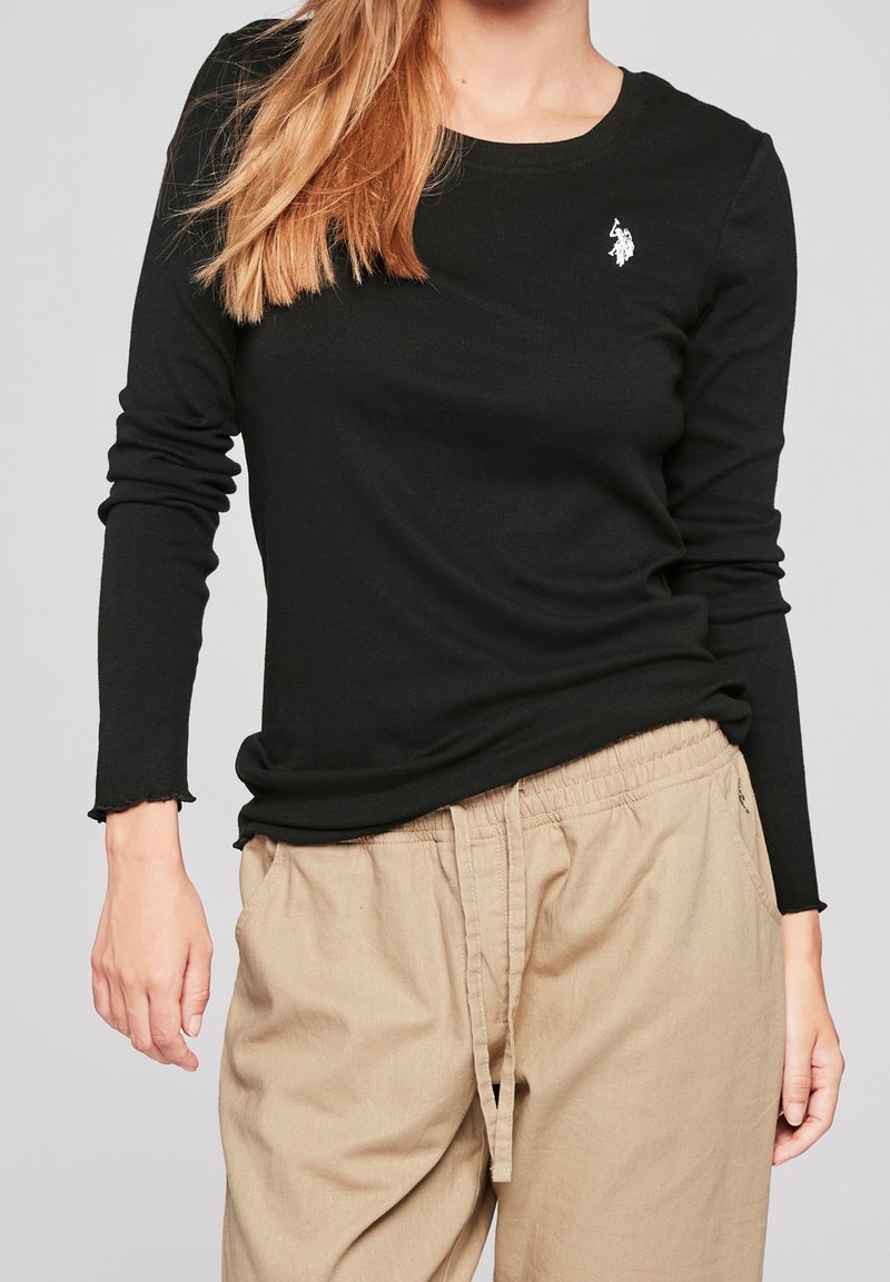 Femme portant un t-shirt noir à manches longues avec un petit logo blanc sur la poitrine et un pantalon beige à cordon sur un fond uni.